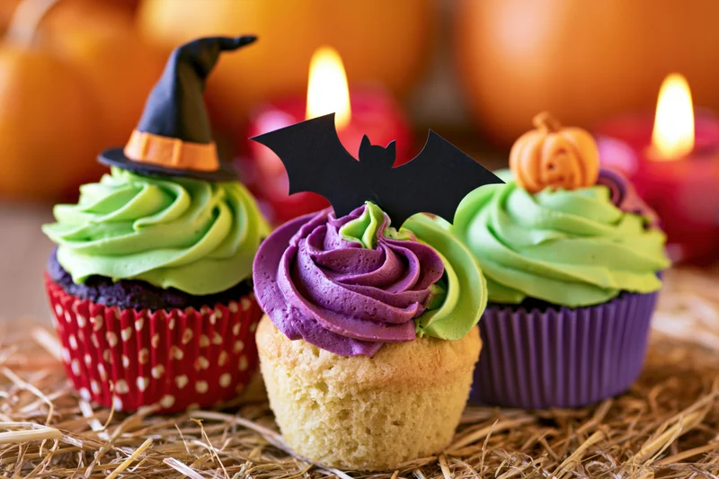 Three halloween-themed cupcakes with witch hat, bat, and pumpkin toppers on straw surface with candles and pumpkins in background