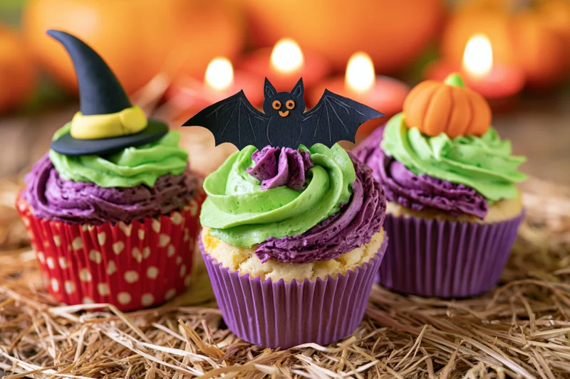Three halloween-themed cupcakes with witch hat, bat, and pumpkin toppers on straw surface