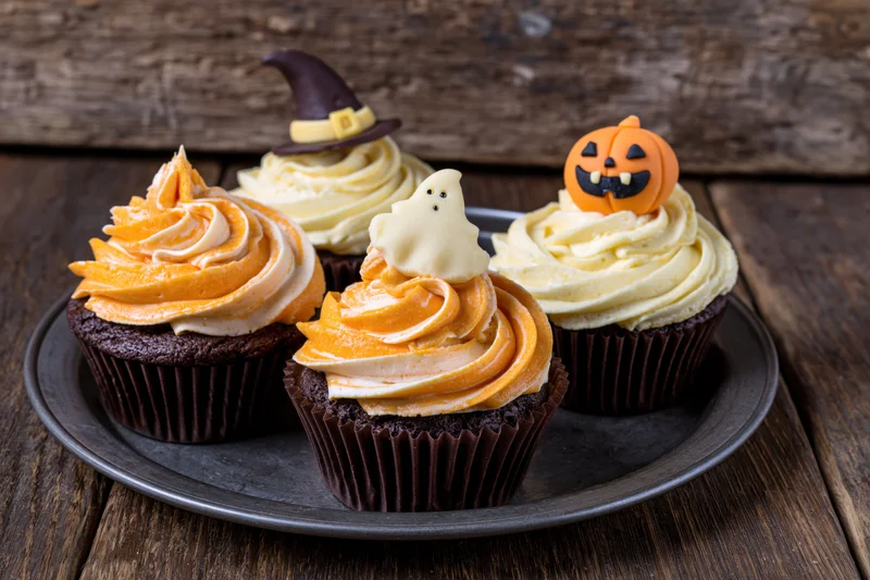 Four halloween chocolate cupcakes with orange and white frosting and spooky toppers on a dark plate on wooden surface
