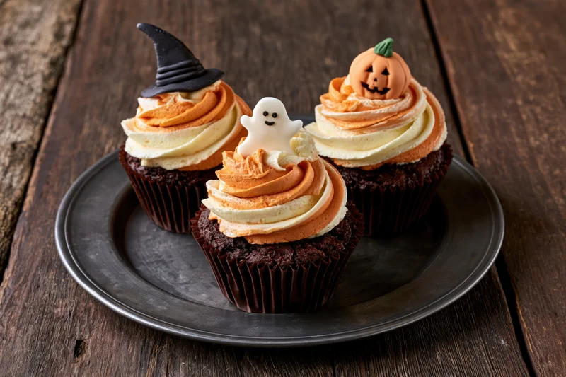 Three halloween chocolate cupcakes with orange and white frosting and spooky decorations on a dark metal plate on wooden table