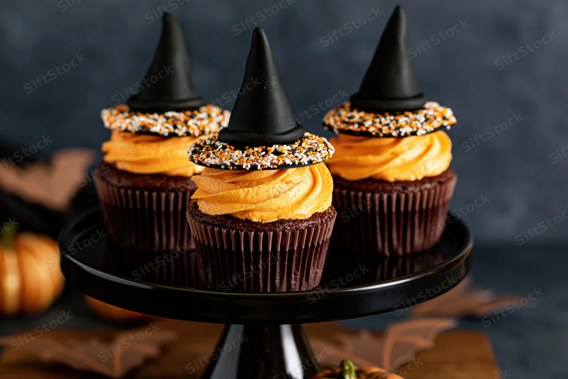 Three halloween cupcakes with orange frosting and witch hats on a black cake stand surrounded by pumpkins and leaves