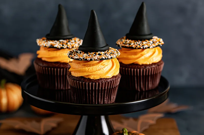 Three halloween cupcakes with orange frosting and witch hats on a black cake stand surrounded by pumpkins and leaves