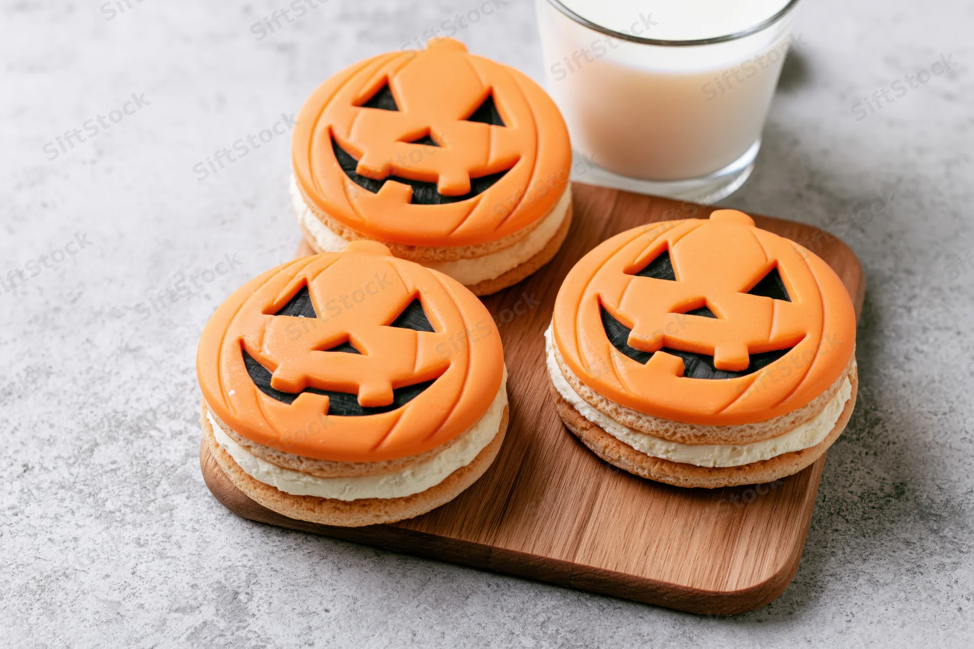 Three halloween jack-o-lantern sandwich cookies with orange frosting and cream filling on a wooden board beside a glass of milk
