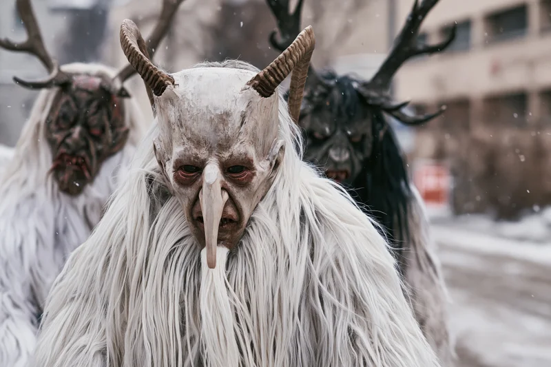 Three krampus performers in traditional alpine costumes with horned masks and fur coats standing in snowy urban setting