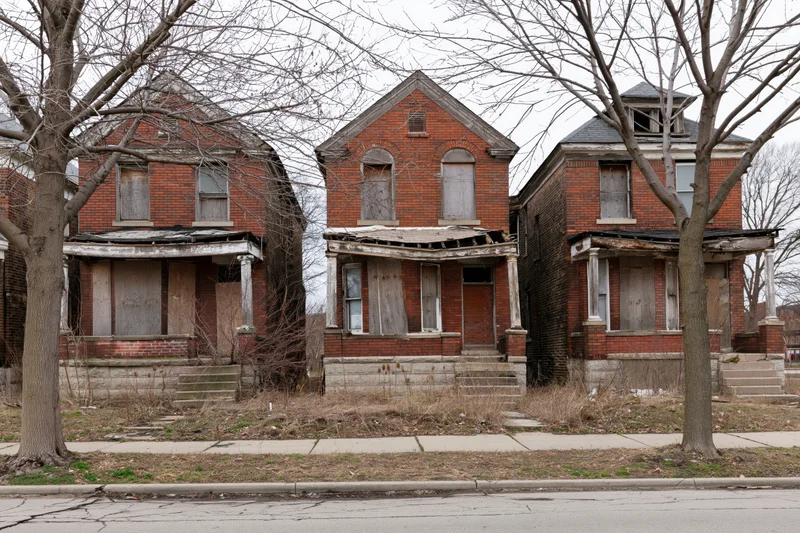 Three abandoned brick row houses with boarded windows and damaged porches on a quiet urban street
