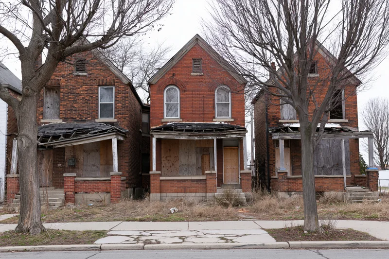 Three abandoned brick row houses with boarded windows and damaged roofs on a quiet urban street