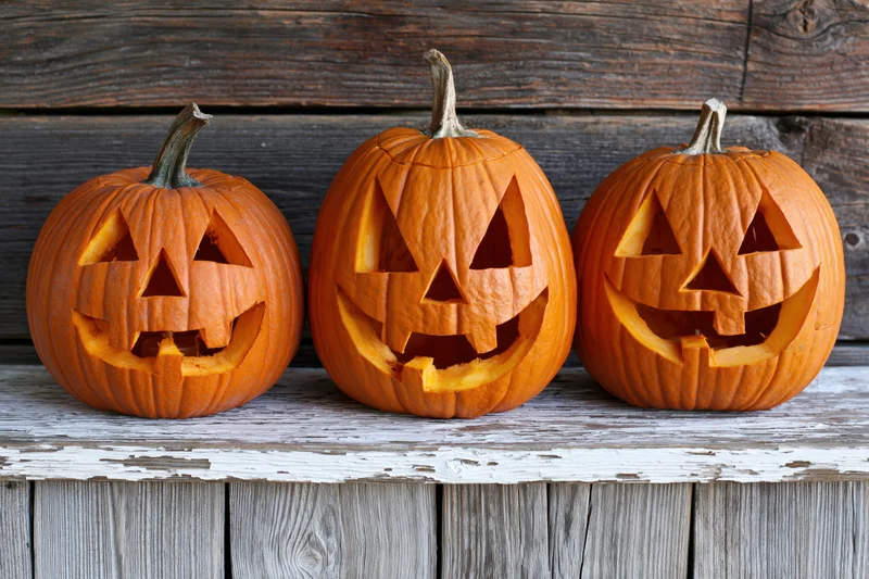 Three carved jack-o-lanterns with triangular eyes and grinning mouths arranged in a row on a weathered wooden shelf