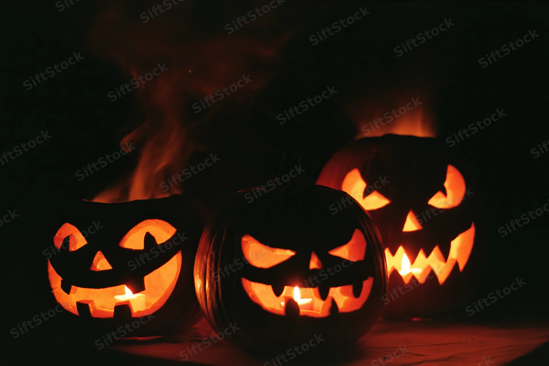 Three carved jack-o-lanterns glowing with candles and smoke at night
