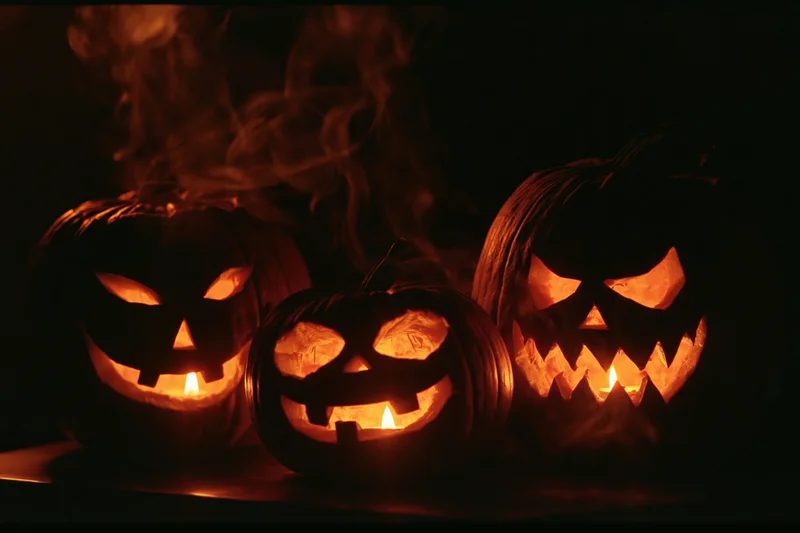 Three carved jack-o-lanterns glowing with candles inside, emitting smoke, arranged in a dark setting