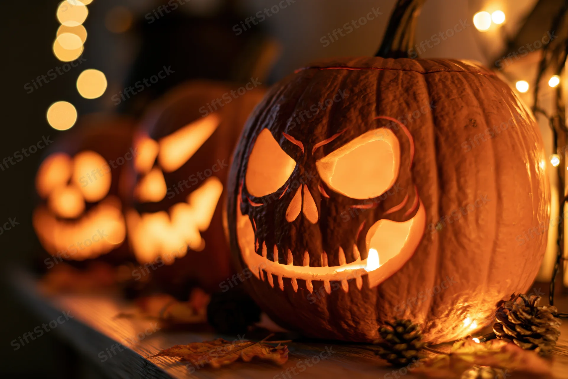 Three carved jack-o-lanterns with skull faces lit from within on a wooden surface surrounded by pinecones and string lights