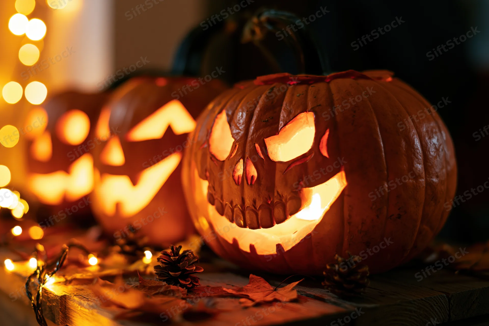 Three carved jack-o-lanterns glowing on a wooden table with autumn leaves and string lights