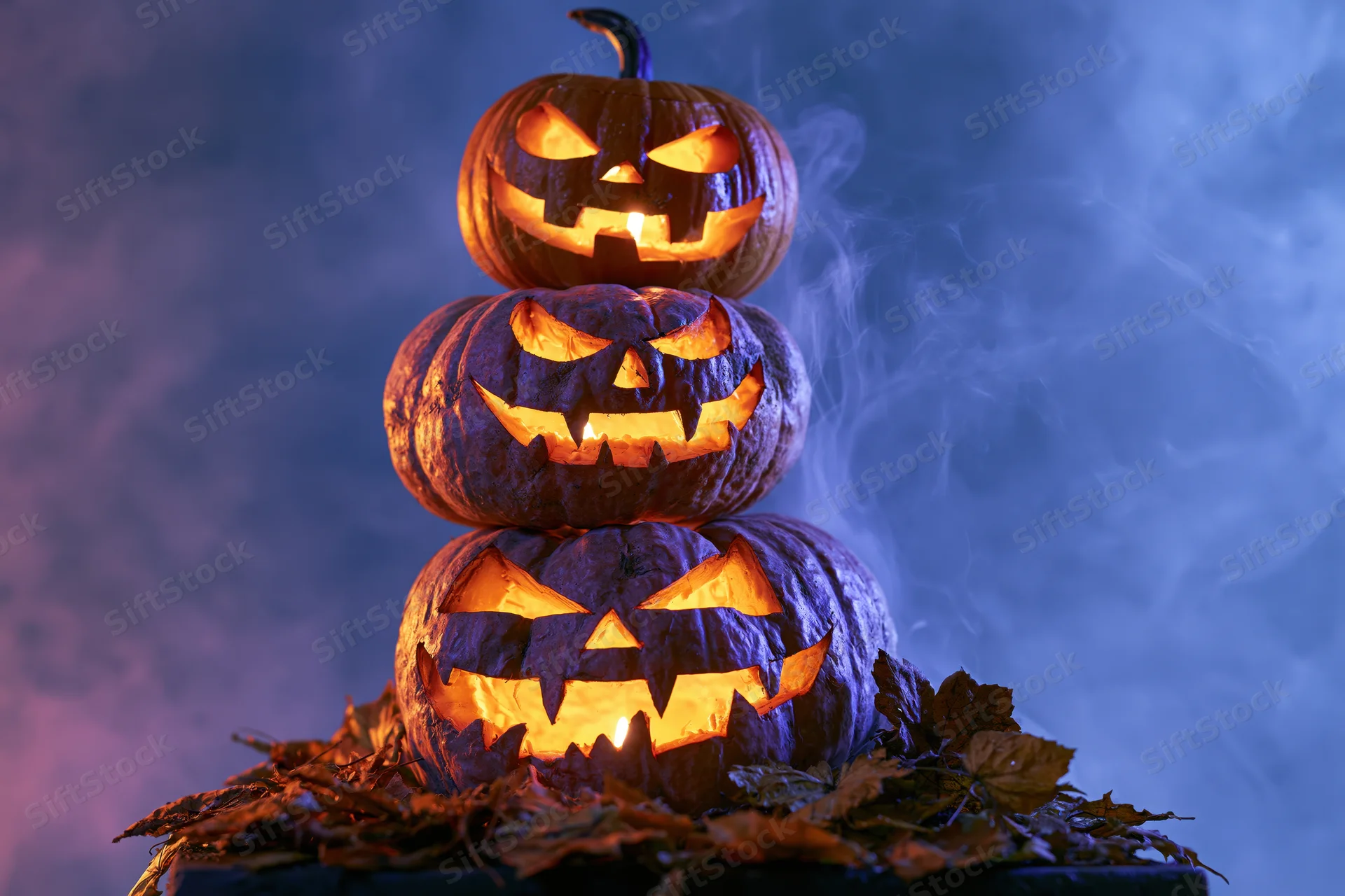 Three carved jack-o-lanterns stacked vertically glowing with candles surrounded by autumn leaves in a smoky blue-lit setting