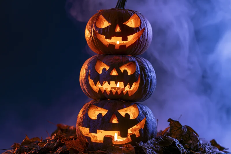 Three carved jack-o-lanterns stacked vertically glowing with candles on a bed of dry leaves in a smoky dark setting