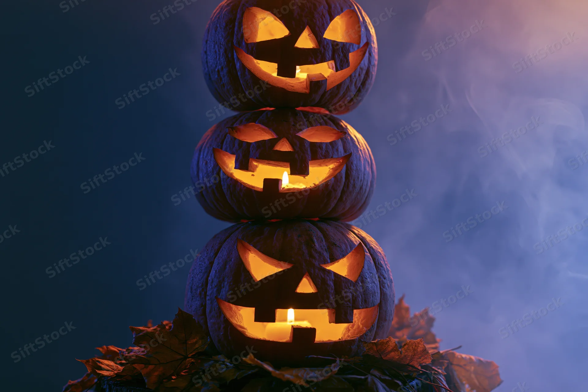 Three carved jack-o-lanterns stacked vertically glowing with candles surrounded by autumn leaves in a smoky dark setting