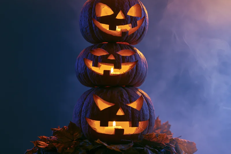 Three carved jack-o-lanterns stacked vertically glowing with candles surrounded by autumn leaves in a smoky dark setting