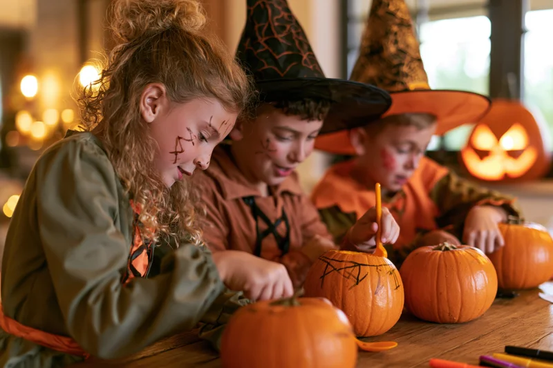 Three children in witch costumes carving pumpkins at a wooden table indoors