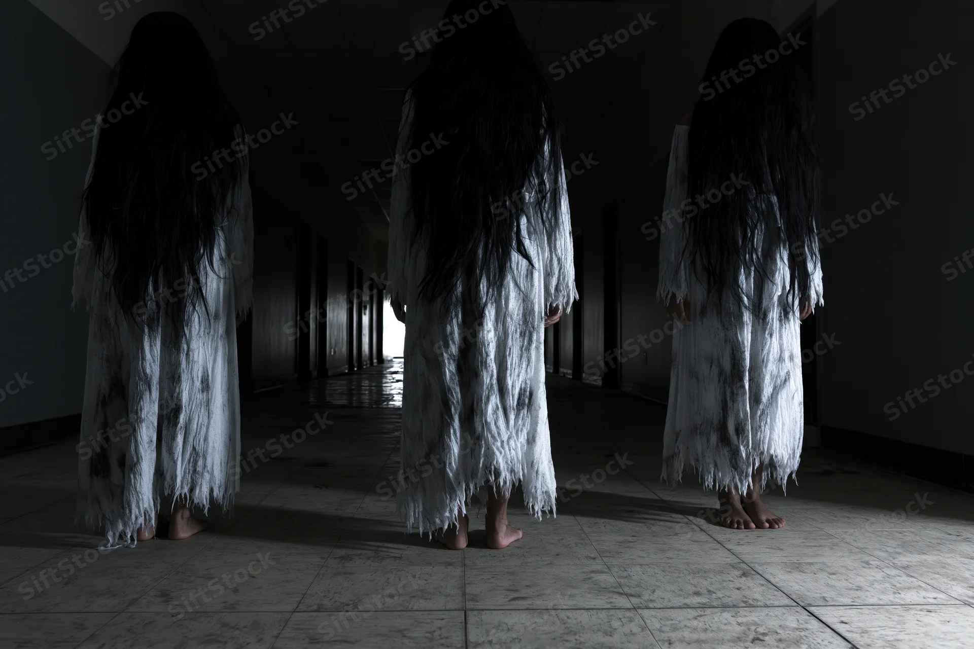 Three women in white gowns with long dark hair standing barefoot in a dim hallway