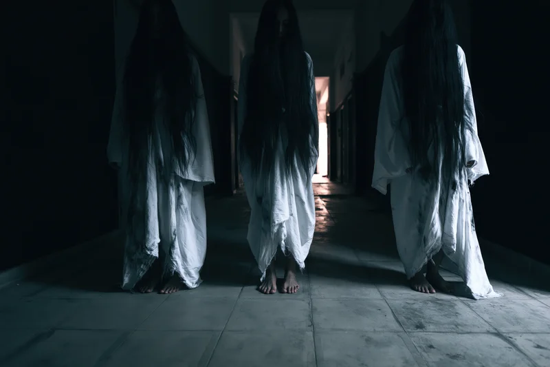 Three ghostly figures in white gowns with long dark hair standing in a dimly lit hallway