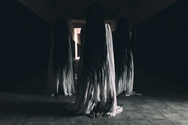 Three women in white gowns with long dark hair obscuring their faces standing in a dim hallway