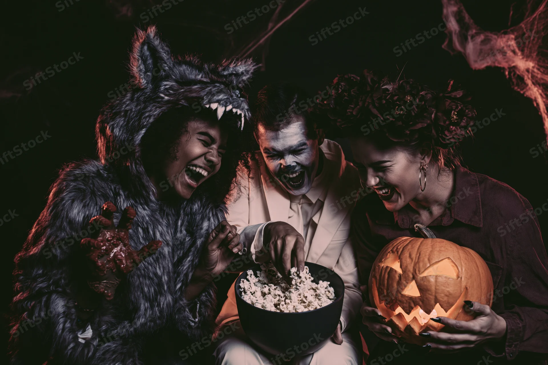 Three friends in halloween costumes laughing together while sharing popcorn and a carved pumpkin indoors