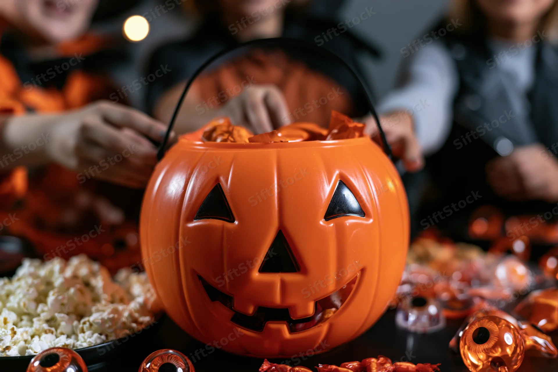 Children in halloween costumes reaching into an orange pumpkin-shaped candy bucket on a table filled with sweets and popcorn