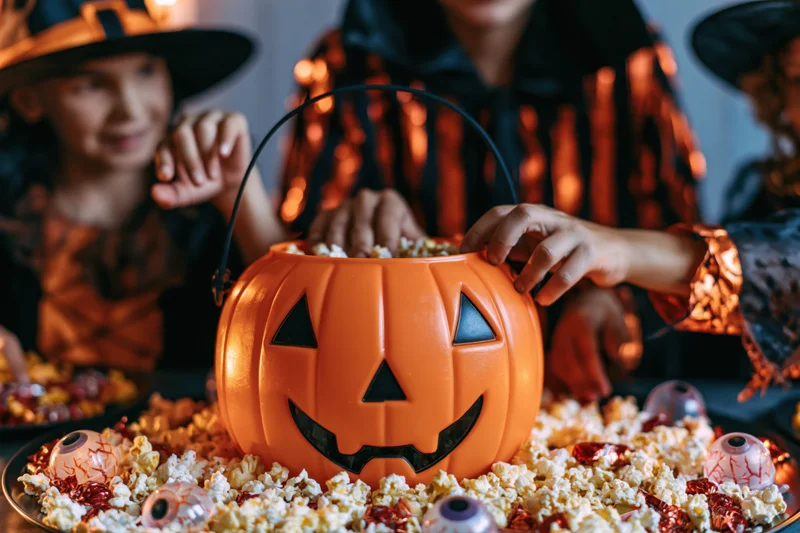 Children in witch costumes reaching into a pumpkin-shaped candy bucket on a table filled with popcorn and candy eyes