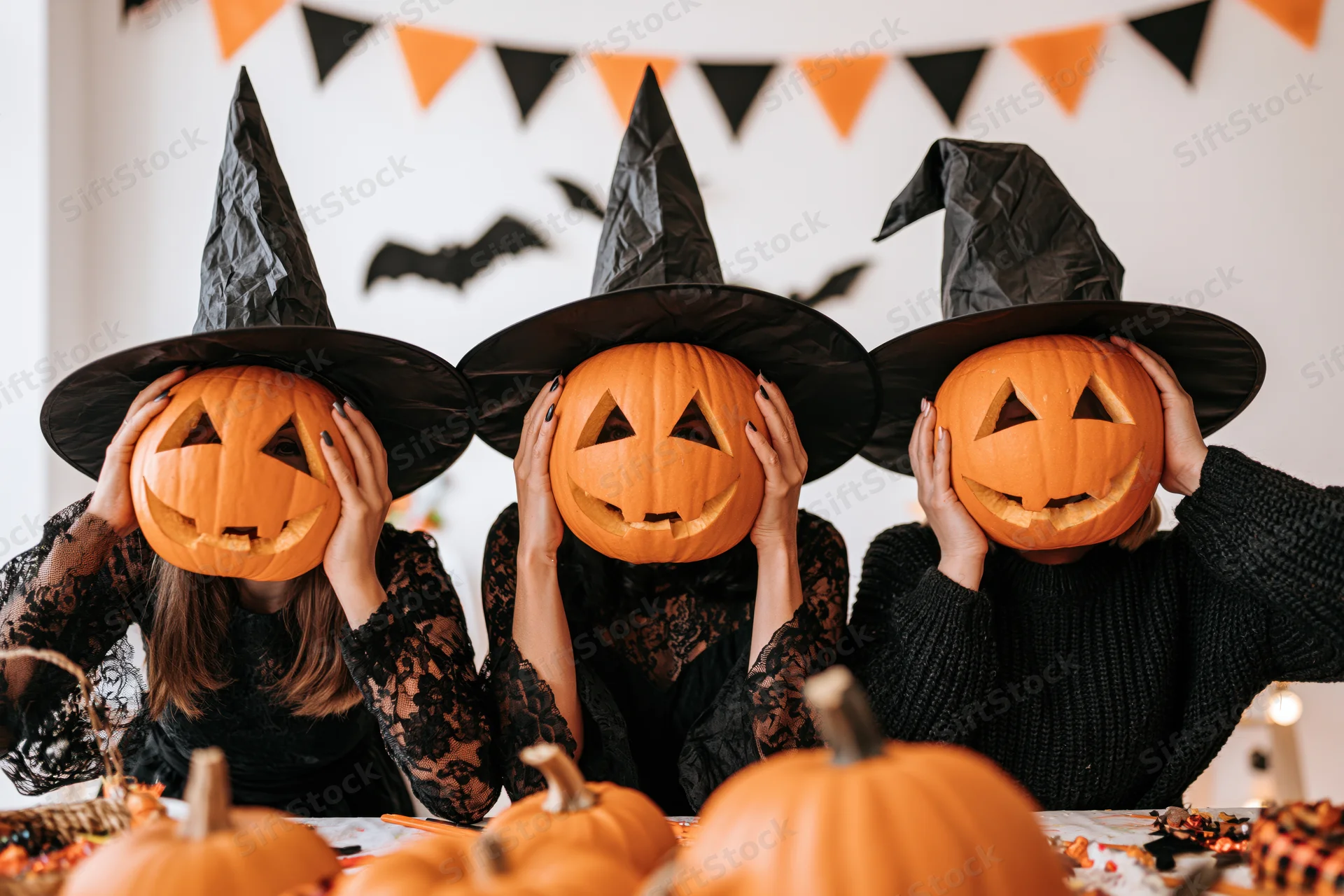 Three women in witch costumes holding carved pumpkins over their faces at a halloween party table