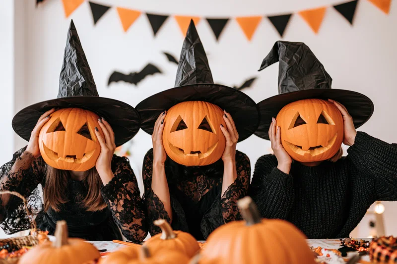 Three women in witch costumes holding carved pumpkins over their faces at a halloween party table
