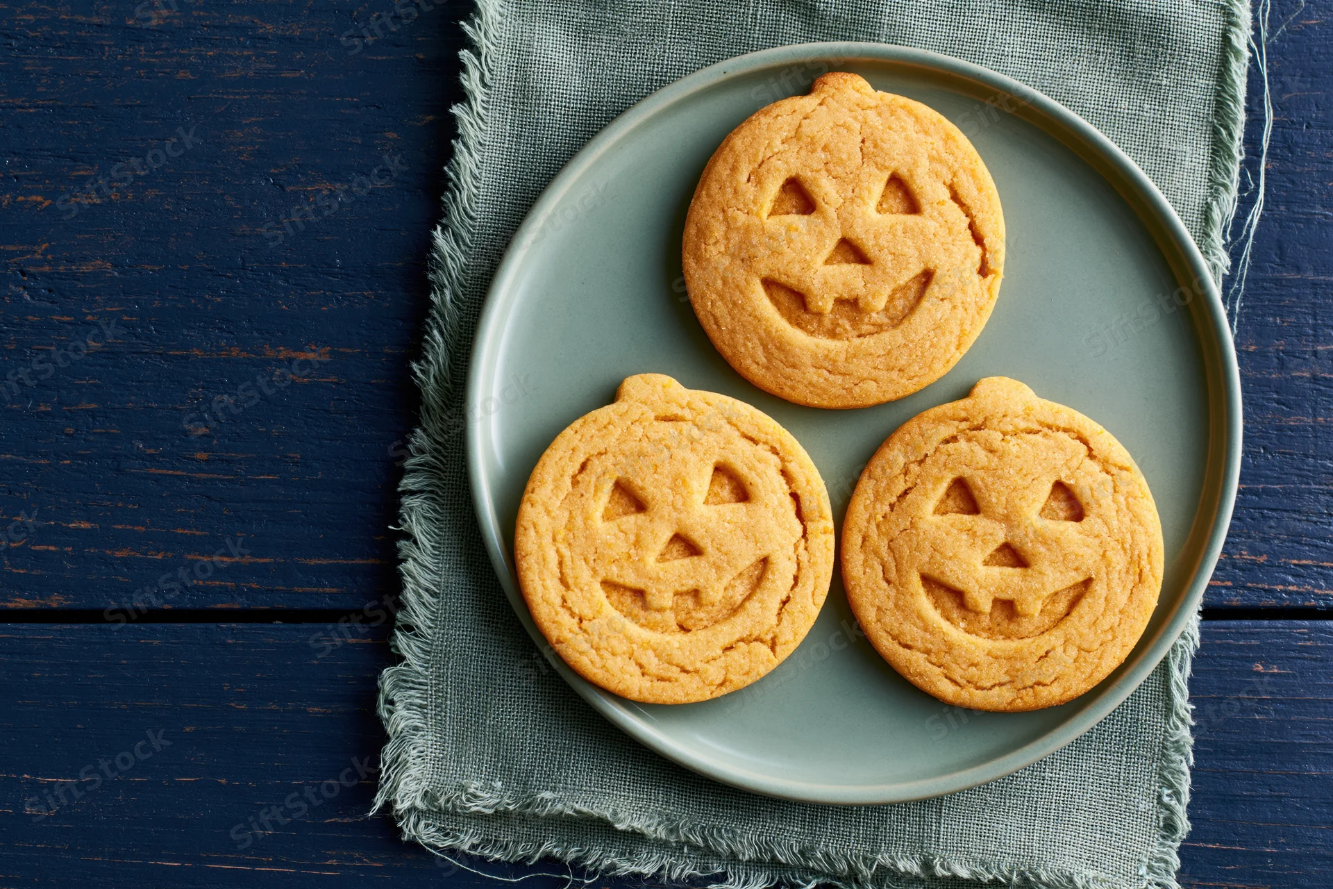 Three jack-o-lantern sugar cookies on a gray plate atop a textured cloth on a blue wooden table