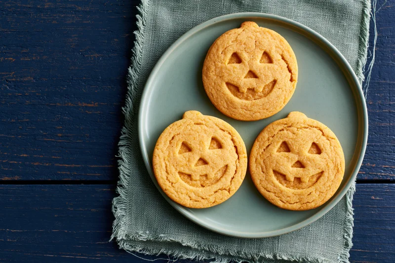 Three jack-o-lantern sugar cookies on a gray plate atop a textured cloth on a blue wooden table