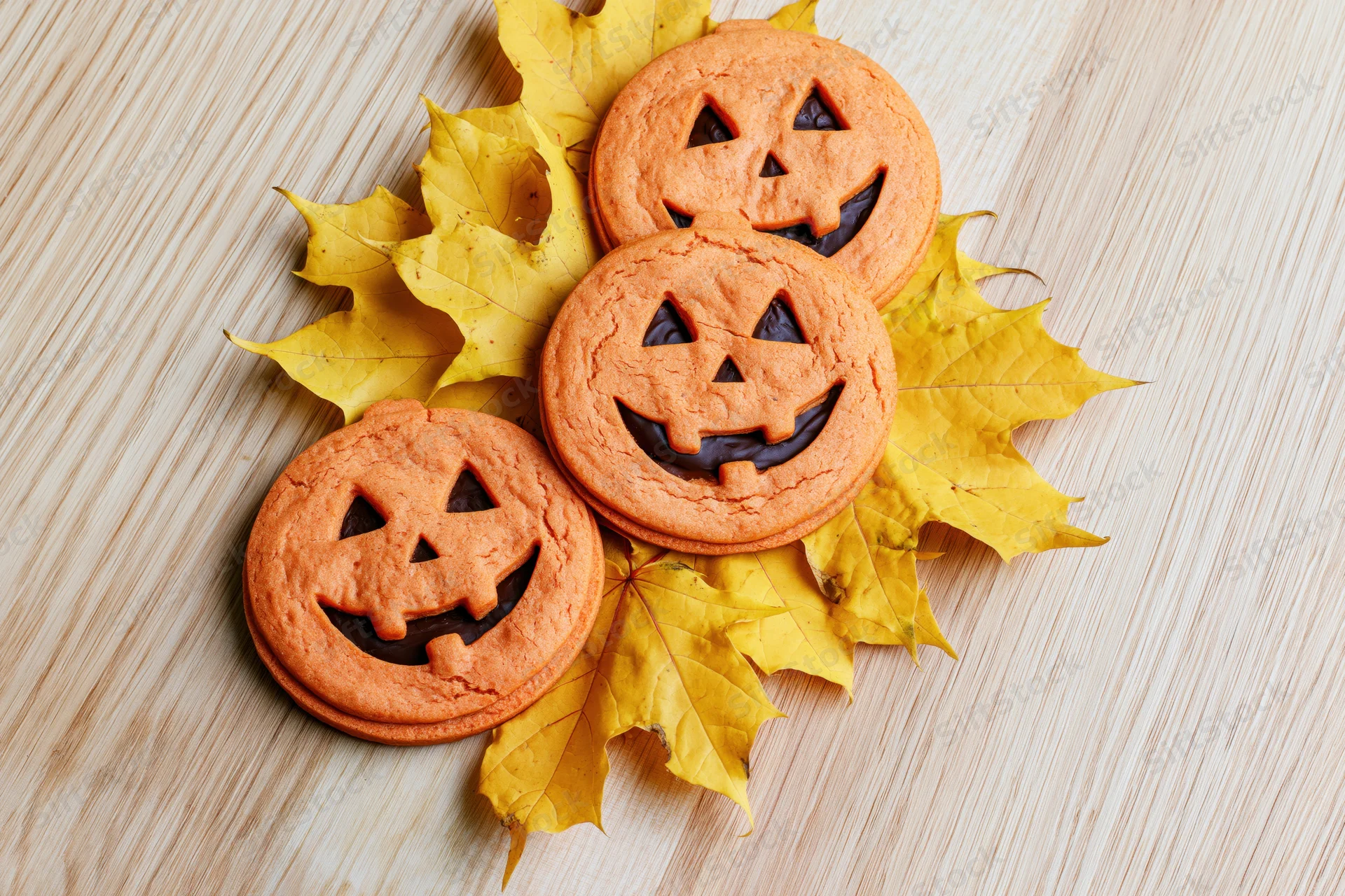 Three orange jack-o-lantern cookies arranged on yellow maple leaves on a wooden surface