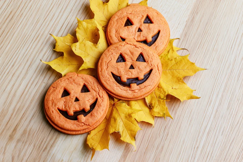 Three orange jack-o-lantern cookies arranged on yellow maple leaves on a wooden surface
