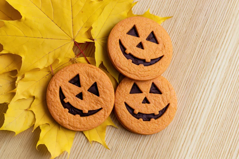 Three round orange jack-o-lantern cookies with chocolate filling on yellow maple leaves and wooden surface