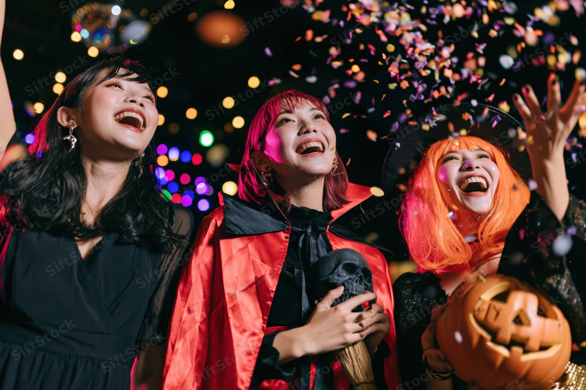 Three young asian women in halloween costumes laughing with confetti and pumpkins at night