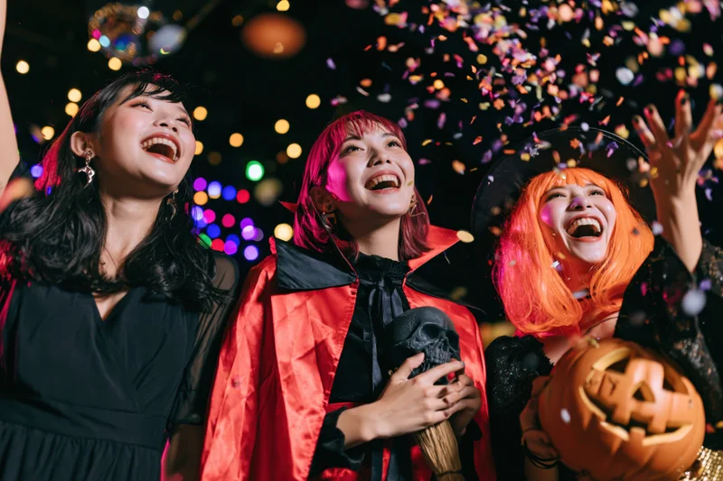 Three young asian women in halloween costumes laughing with confetti and pumpkins at night