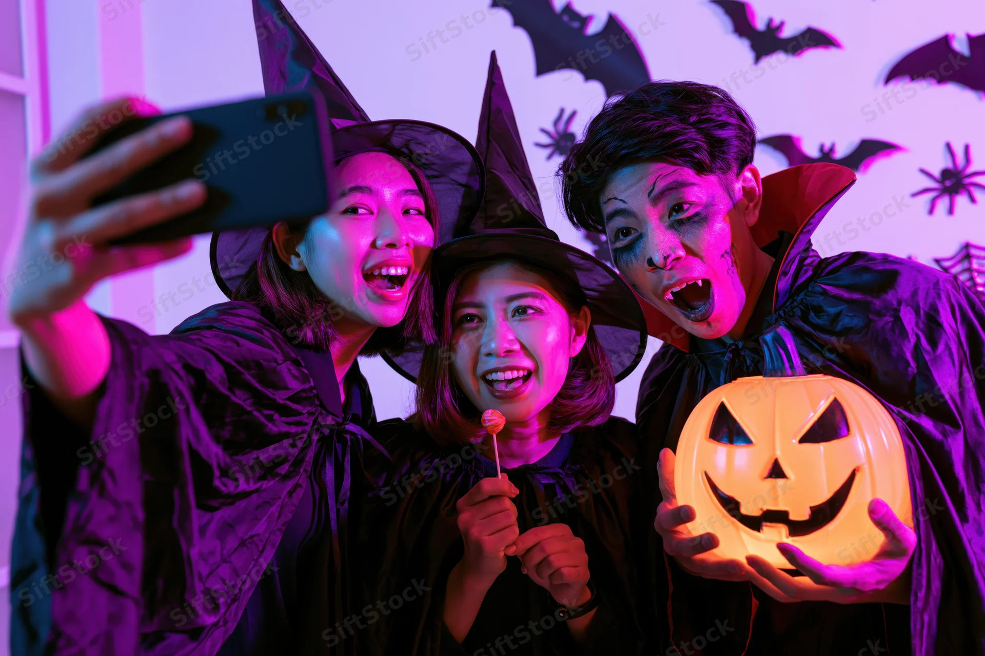 Three young asian adults in halloween costumes taking a selfie indoors with glowing pumpkin and decorations