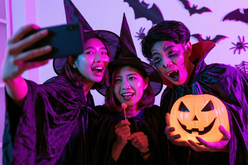 Three young asian adults in halloween costumes taking a selfie indoors with glowing pumpkin and decorations