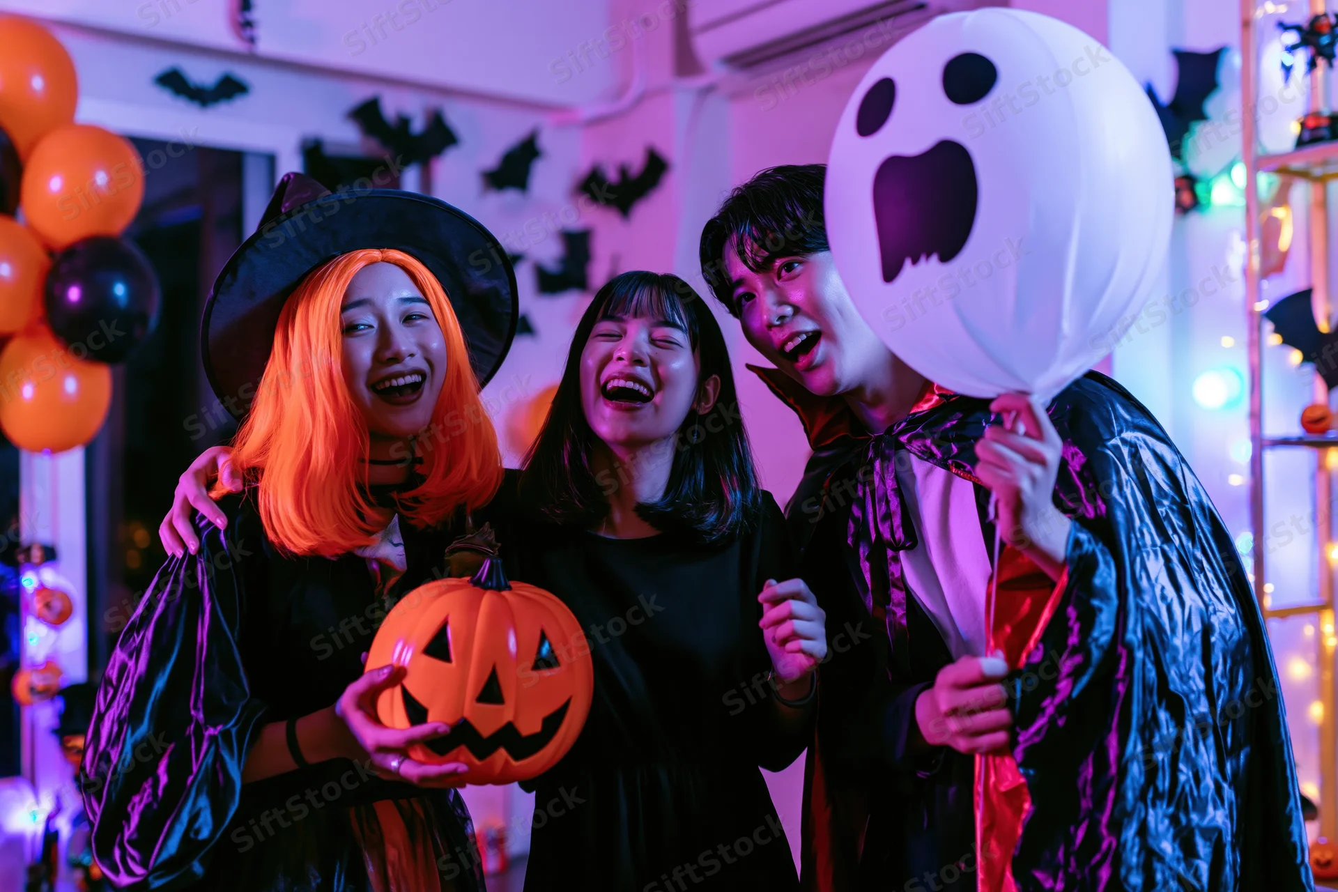 Three young asian friends laughing together at a halloween party in costumes with pumpkins and balloons