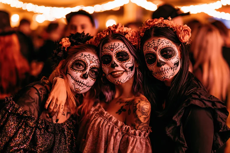 Three young women with sugar skull face paint embracing at a festive outdoor party under string lights