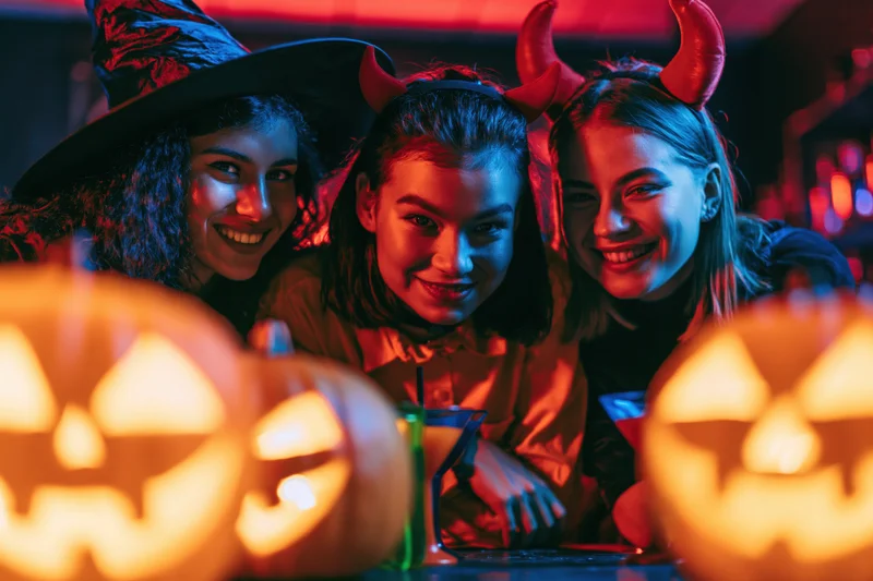 Three young women in halloween costumes smiling at a bar with jack-o-lanterns in the foreground