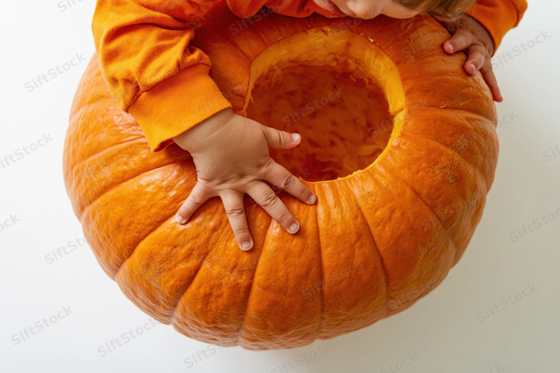 Toddler reaching into a carved pumpkin with hands on its sides, top view on white background