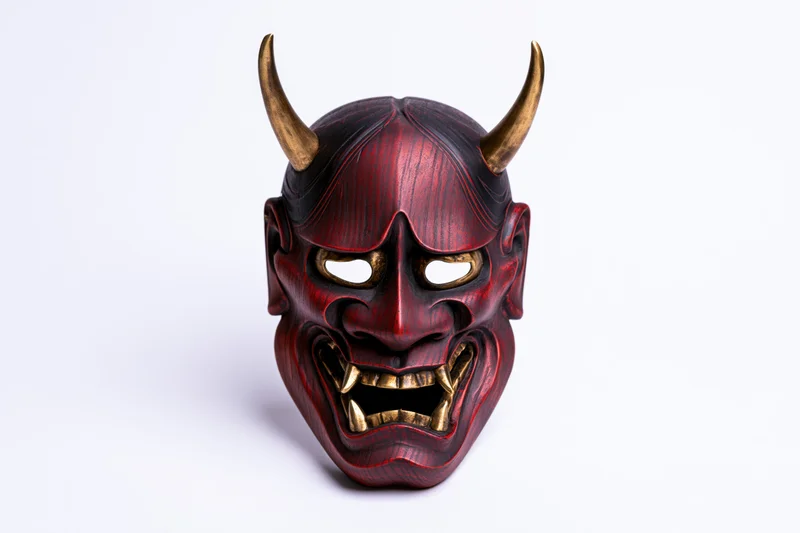 Traditional japanese hannya noh theater mask with red lacquered wood and gold horns displayed face-on against white background