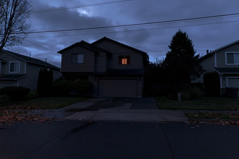 Two-story suburban house at night with a single dimly lit window and scattered autumn leaves on the driveway and sidewalk