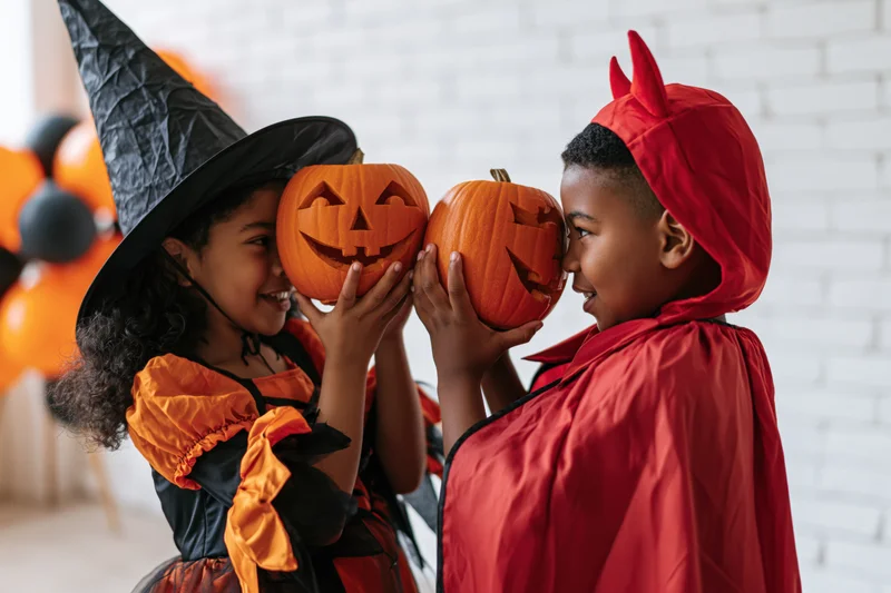 Two black children in halloween costumes holding carved pumpkins facing each other indoors
