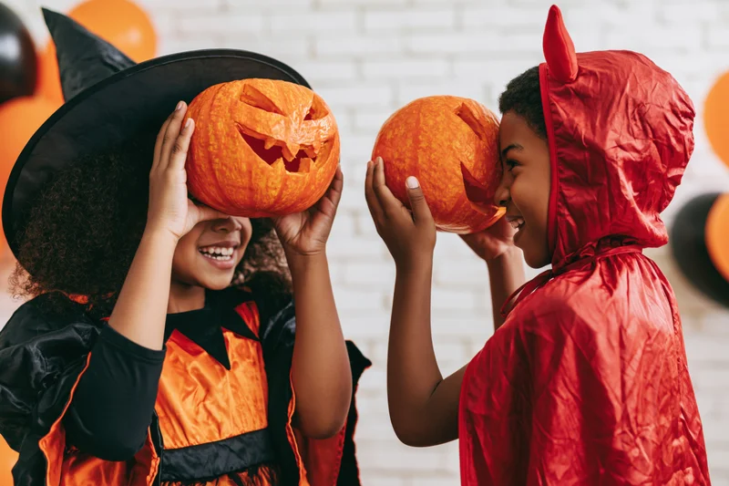 Two black children in halloween costumes holding carved pumpkins over their faces, smiling at each other indoors