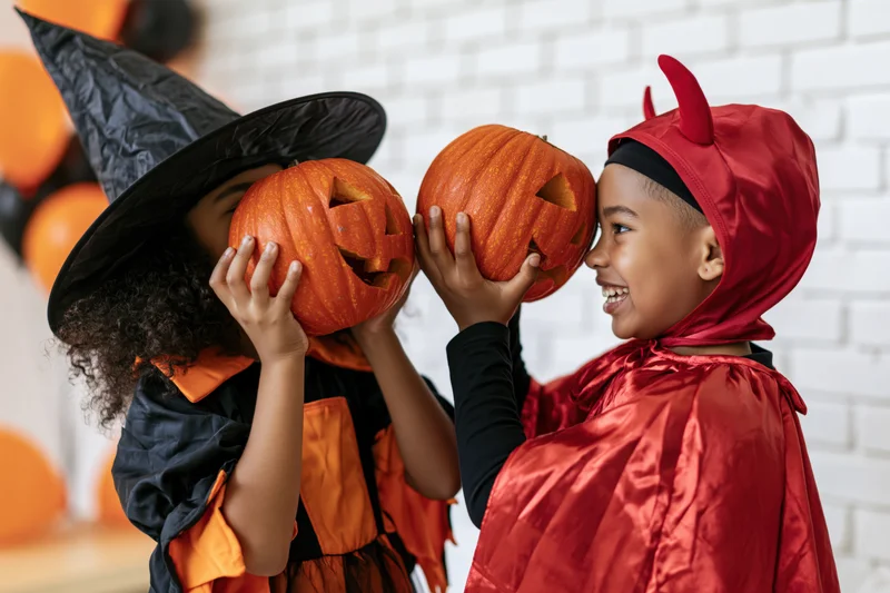 Two children in halloween costumes holding pumpkins and smiling at each other indoors