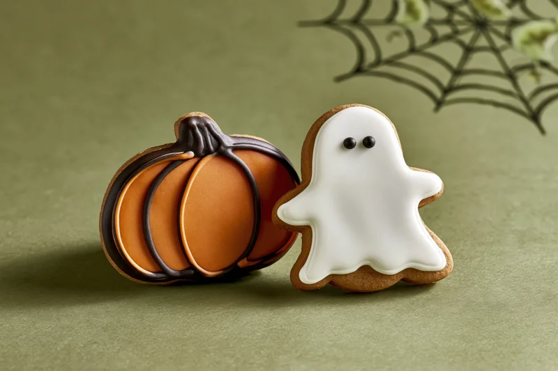 Two halloween gingerbread cookies shaped as a pumpkin and a ghost on a green surface with a spiderweb decoration