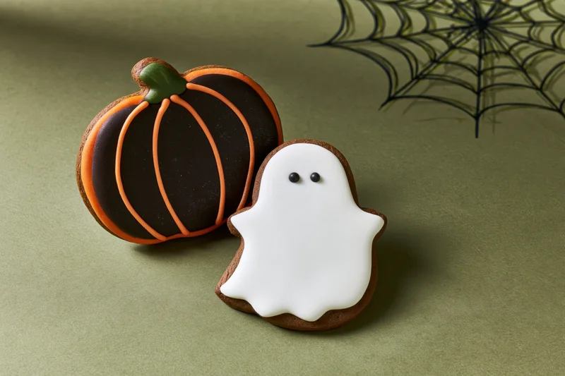 Two halloween gingerbread cookies shaped as a pumpkin and a ghost on a green surface with a spiderweb decoration