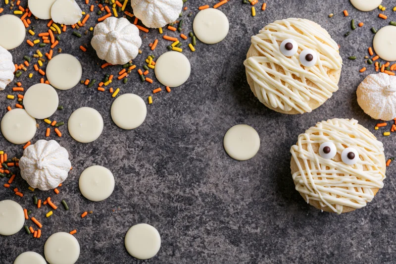Two halloween mummy cupcakes decorated with white icing drizzle and candy eyes on dark surface with sprinkles and white chocolate discs around