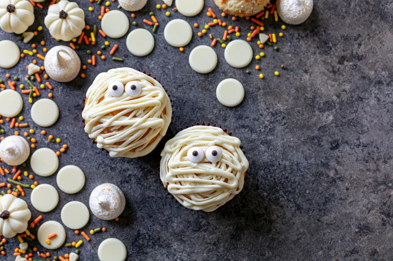Two halloween mummy cupcakes decorated with white icing drizzle on dark surface with sprinkles and candy around